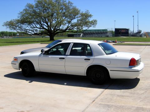 2000 Ford Crown Victoria - A white 2000 Ford Crown Victoria sedan is parked on a concrete lot next to another vehicle, with a large tree and stadium in the background.