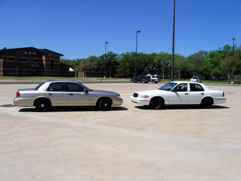 2000 Ford Crown Victoria - Two Ford Crown Victorias are parked in an open lot on a sunny day, with a brick building in the background.