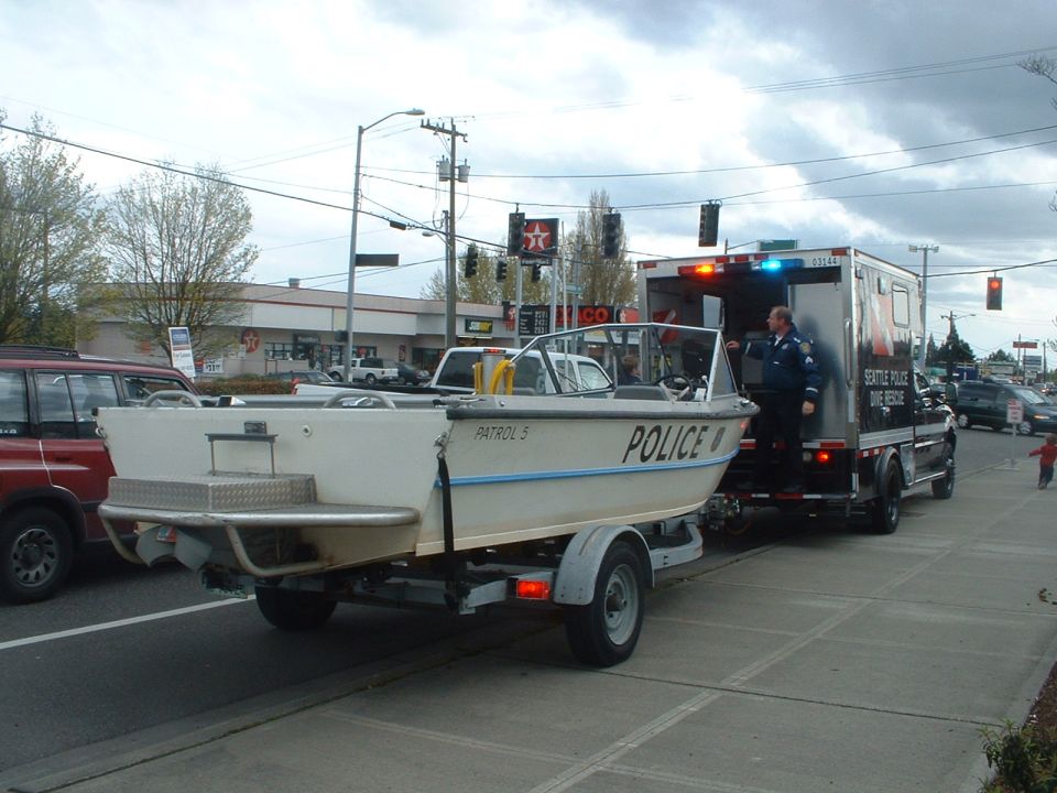 White police boat on a trailer, towed by a dark truck with emergency lights