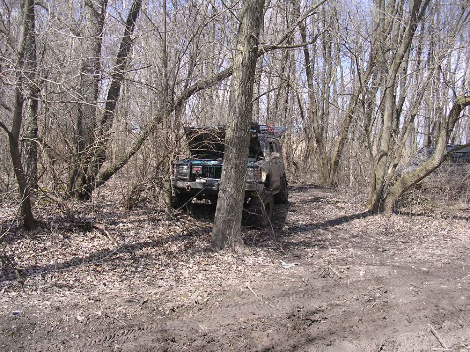 Dark green Jeep Cherokee with hood open, parked in a wooded area with bare trees
