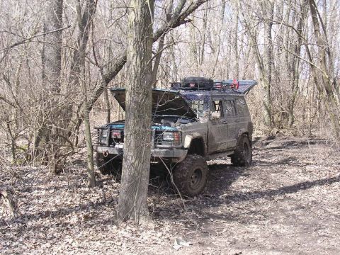 1995 Jeep Cherokee - A mud-covered 1995 Jeep Cherokee sits in a wooded area with its hood and rear hatch open.