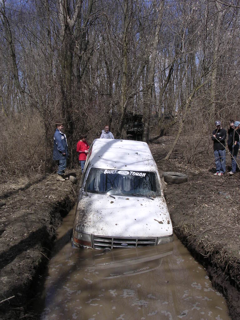 White Ford truck stuck in a muddy ditch, front view, with 'Built Ford Tough' sticker on windshield