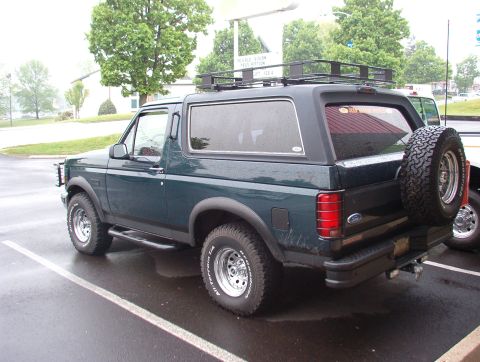 1993 Ford Bronco - A 1993 Ford Bronco is seen from a three-quarter rear view, parked in a wet lot with a roof rack and spare tire.
