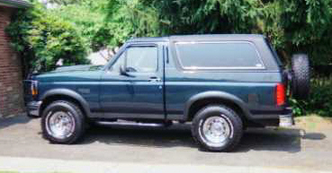 1993 Ford Bronco - A dark green 1993 Ford Bronco is parked outside next to some trees and a brick wall.