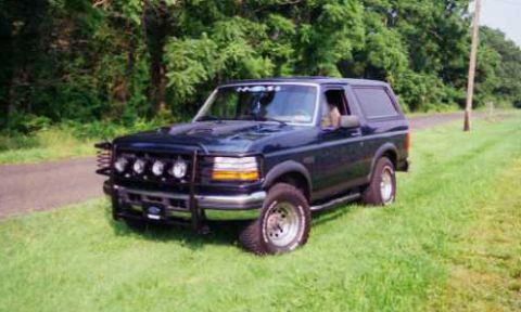 1993 Ford Bronco - A dark blue 1993 Ford Bronco is parked on a grassy area next to a dirt road, a prominent brush guard with multiple lights.