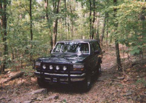 1993 Ford Bronco - A dark green 1993 Ford Bronco navigates a wooded trail, equipped with a bull bar and auxiliary lights.