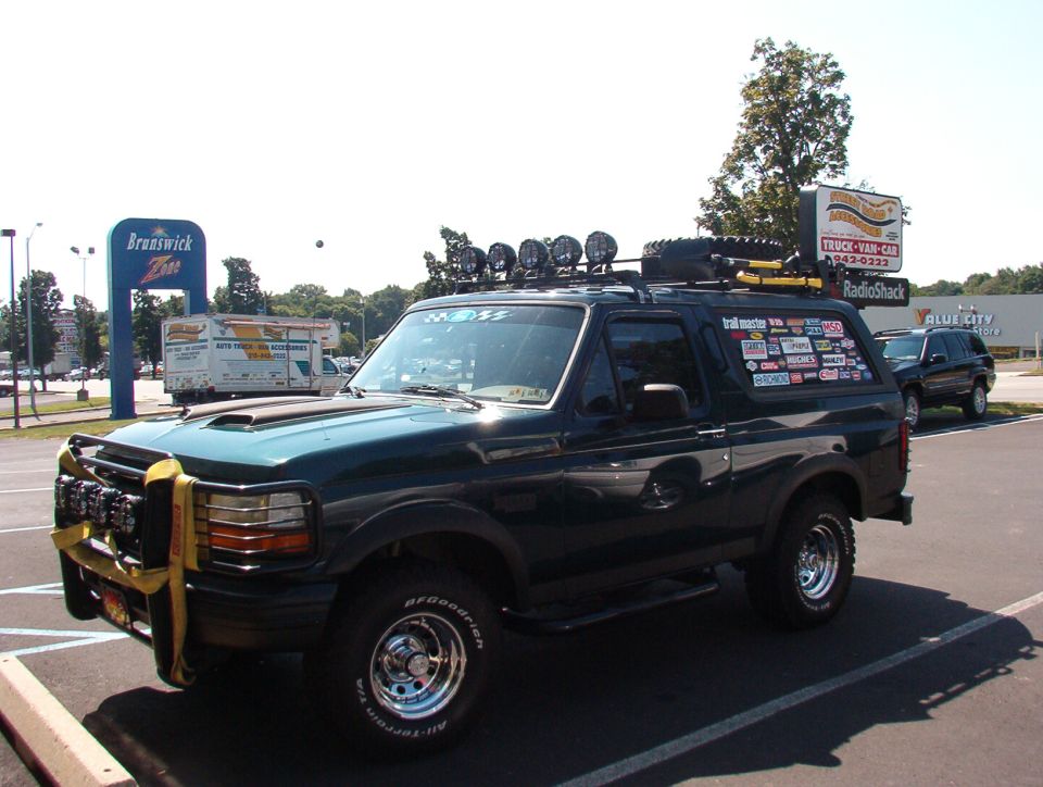 Dark green Ford Bronco with roof rack, spare tire, and many stickers on the rear window. Another SUV is parked nearby