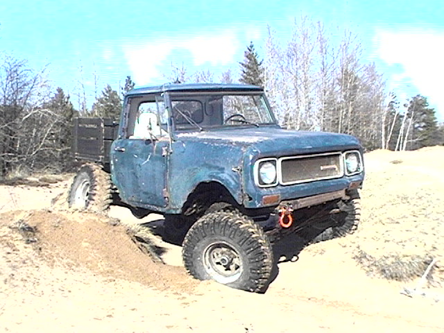 Faded blue lifted International Scout on big tires flexing over a sand dune.