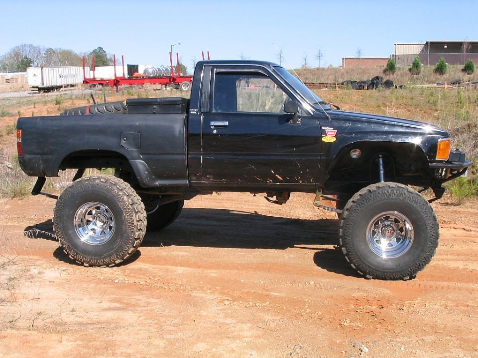 Lifted black pickup truck with large tires, parked on dirt. Has a toolbox in the bed
