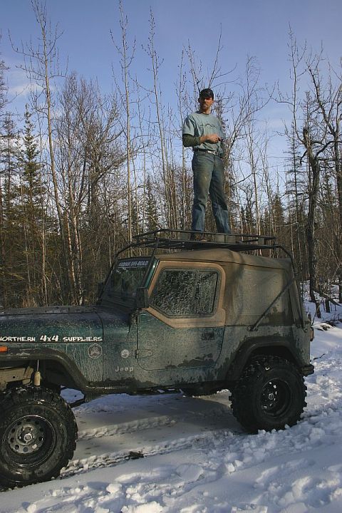 2003 Jeep TJ - A man stands atop a modified Jeep Wrangler, parked in a snow-covered, wooded landscape.