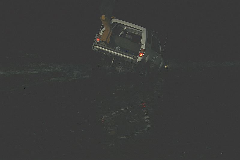 White SUV stuck in dark water, rear view with open trunk and person standing in it