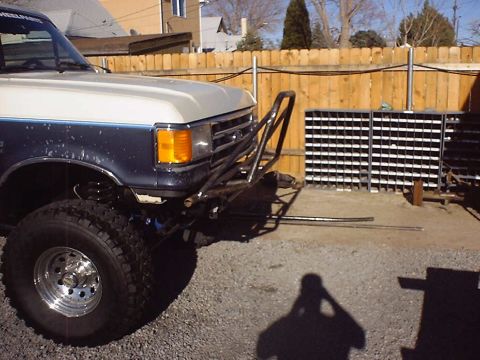 1988 Ford Bronco - Front three-quarter view of a lifted blue-and-white two-tone Ford truck on chrome eight-lug wheels and tall mud-terrain tires, with a bare-…