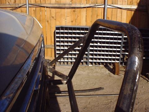 1988 Ford Bronco - A custom roll cage is welded to the side of a vehicle, with a storage rack and wooden fence visible behind it.
