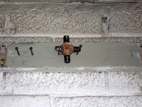 Plumbing cross fitting attached to a wooden board with nails, mounted on a brick wall