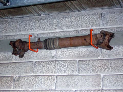 1995 Ford Bronco - A rusty driveshaft is hung on orange hooks against a textured brick wall.