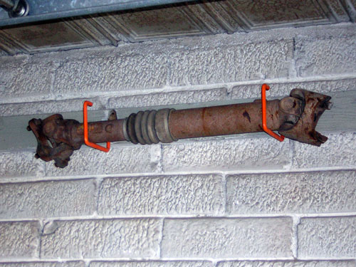 Rusty driveshaft with a rubber boot, mounted on orange hooks against a brick wall