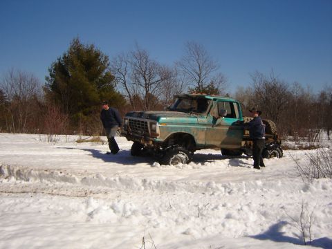 1978 Chevrolet C/K Pickup - A teal pickup truck with oversized tires is stuck in a snowy field, with two people nearby.
