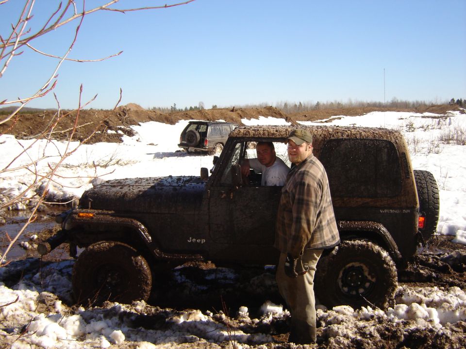 Muddy Jeep Wrangler with two men inside, stuck in snow and mud. Another SUV is visible in the background