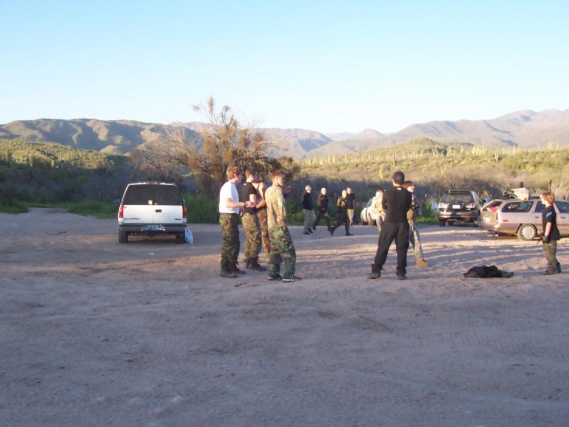 White full-size SUV parked in a desert dirt lot with people in camo standing nearby and other cars at right