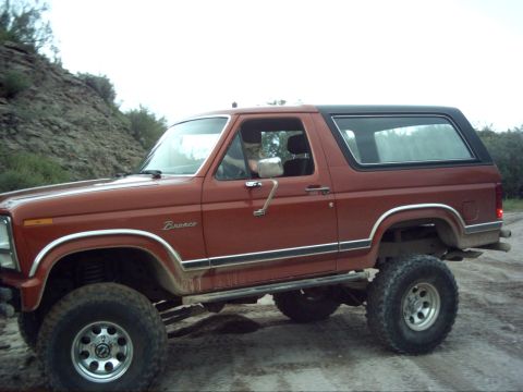 1988 Ford Bronco - A lifted red Ford Bronco with a black top is parked on a dirt road with a rocky hill in the background.