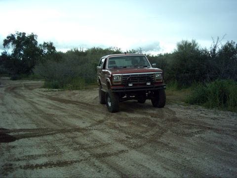 1988 Ford Bronco - This lifted SUV is seen off-road on a dusty track with brush in the background.