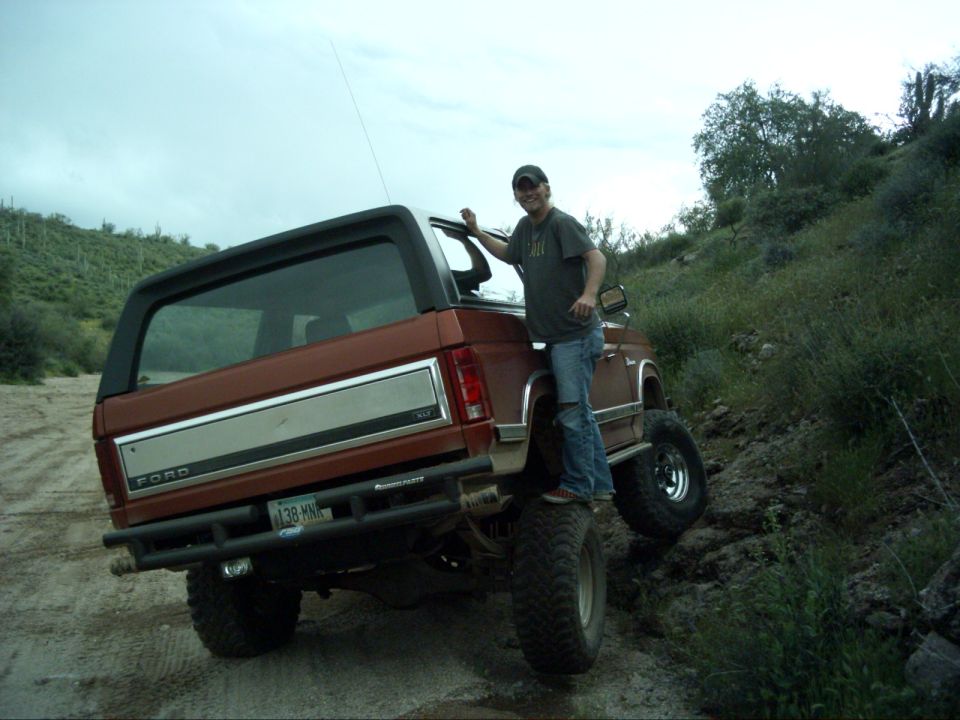 Man in cap stands on the tire of a lifted copper-brown Bronco on a desert hillside, rear view