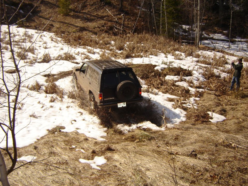 Muddy two-door SUV with rear tire carrier crossing a snowy wooded trail rut