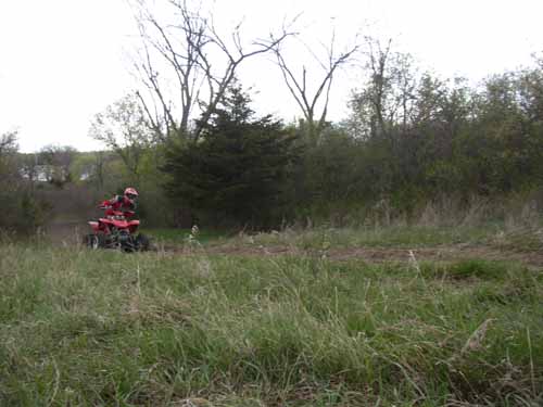 Rider on a red ATV crossing a grassy field with trees in the background