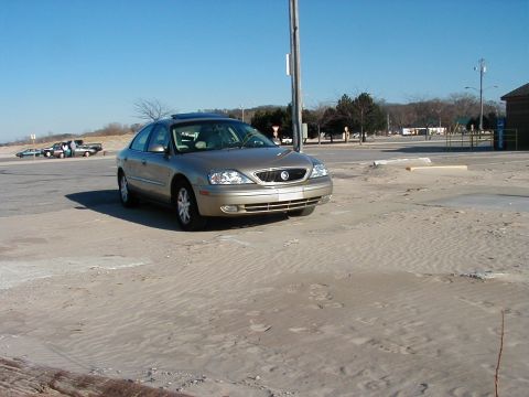 1997 Ford Taurus - A gold sedan is parked on a sandy lot under a clear blue sky. The vehicle's front end is visible, showing its grille and headlights.