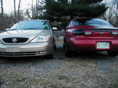 1997 Ford Taurus - Two sedans, a gold one and a red one, are parked next to each other on a gravel lot.
