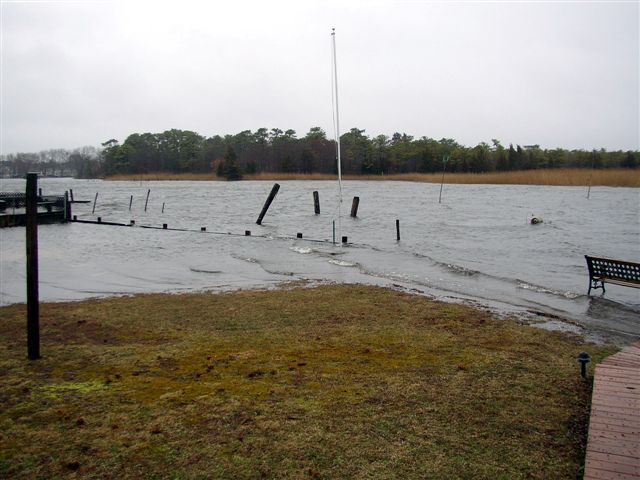 Flooded waterfront with submerged bulkhead, pilings and a bench under gray storm sky