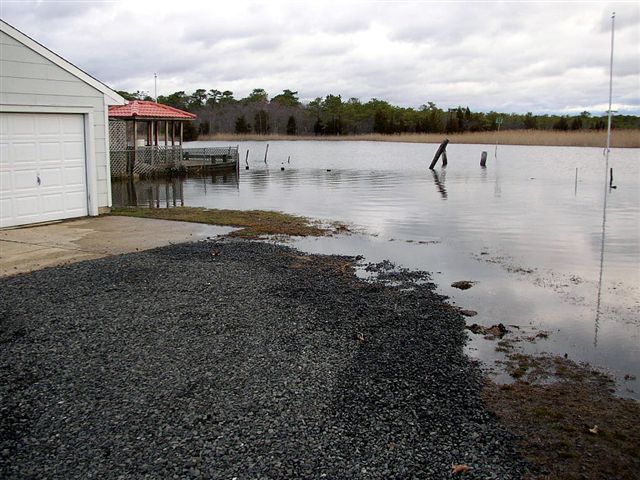 Storm floodwater over a gravel driveway beside a white garage and waterfront gazebo