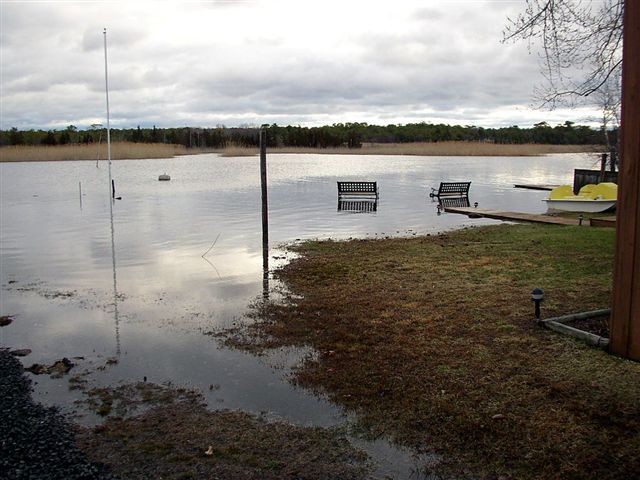 Flooded yard with two benches partially submerged in water. A yellow boat is docked on the right