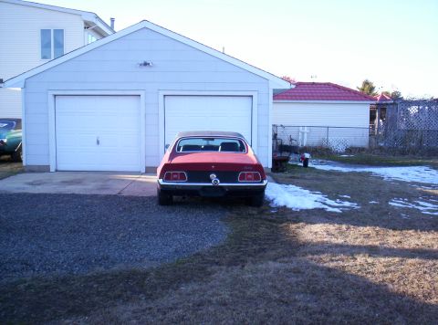 1995 Ford Bronco - Rear view of a red 1970s Ford Mustang coupe with a tan vinyl roof, parked nose-in at a white two-door detached garage on a gravel driveway.…