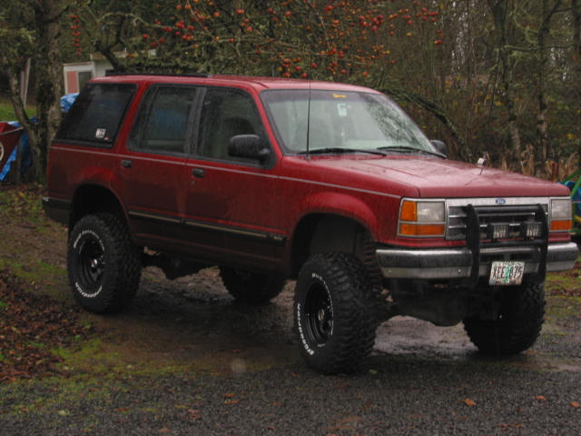 Lifted dark red Ford Explorer SUV with mud tires and brush guard parked on wet gravel driveway