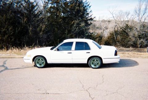 2000 Ford Crown Victoria - A white 2000 Ford Crown Victoria sedan is viewed from the side, parked on a road with trees in the background.