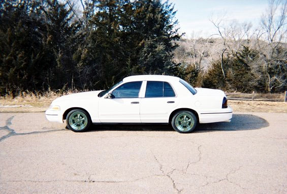 White Ford Crown Victoria sedan side profile on cracked asphalt with bare winter trees