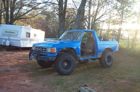 1987 Ford Bronco - A blue pickup truck sits in a grassy area next to a camper, appearing to be undergoing some modifications.