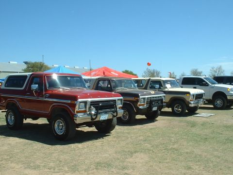 1978 Ford Bronco - A row of SUVs, including a red 1978 Ford Bronco, are parked on a grassy field under a clear blue sky.