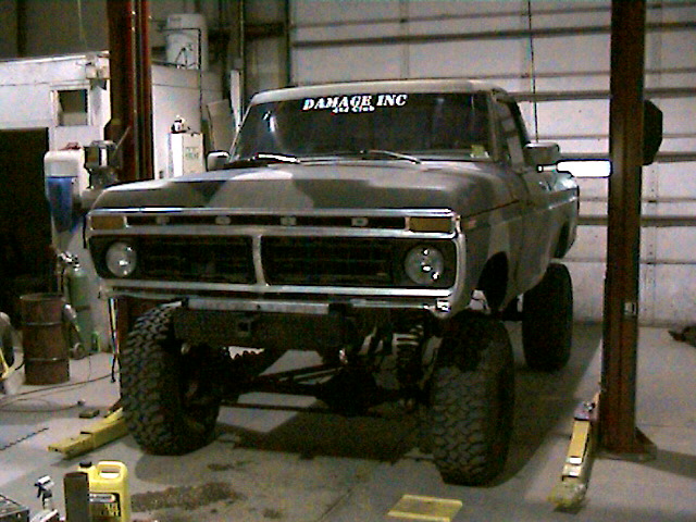 Lifted 1970s Ford pickup on a shop lift, gray primer, 'DAMAGE INC' lettered on windshield