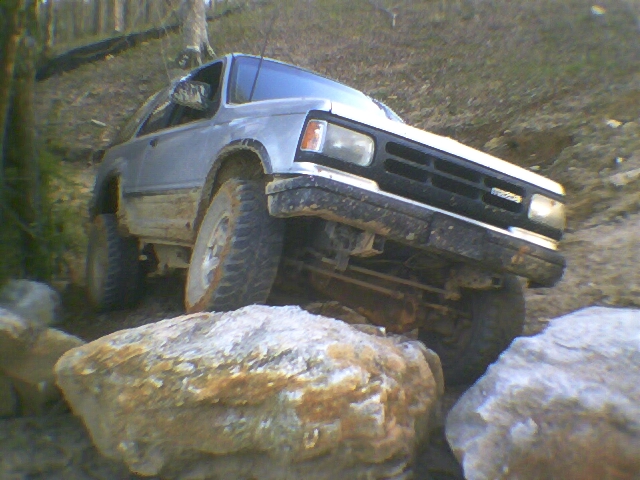 Silver two-door SUV on mud-terrain tires crawling over boulders on a wooded trail