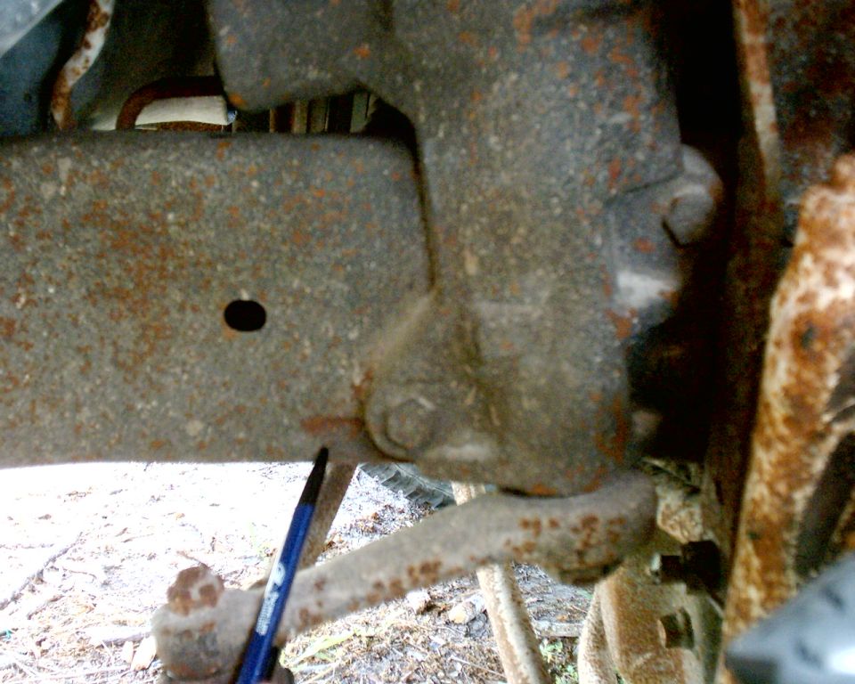 Close-up of heavily rusted vehicle frame and suspension parts, with a blue pen pointing to a bolt
