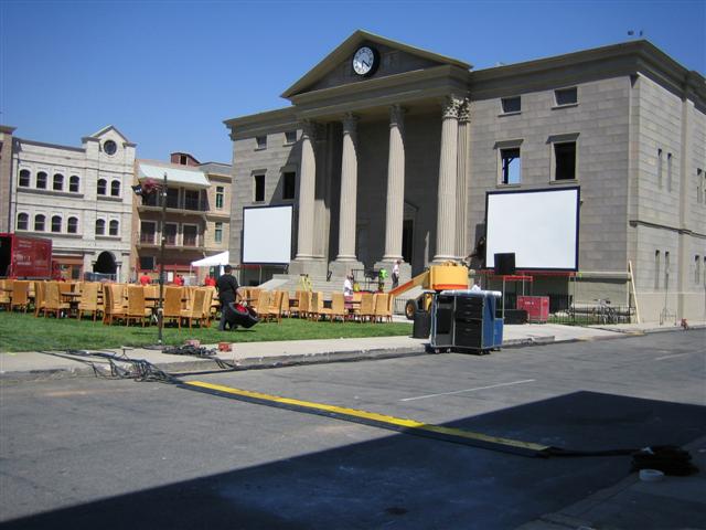 Film set with a courthouse-like building, chairs on grass, and two large screens