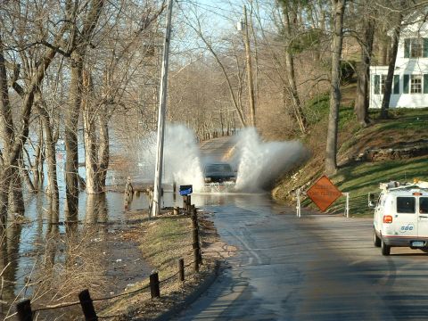 1993 Ford Bronco - A dark SUV drives through floodwaters on a rural road, with a white van and a "Road Flooding" sign visible.
