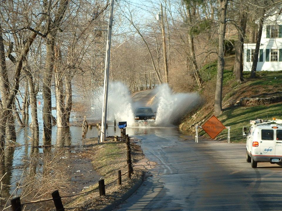 Dark SUV driving through floodwaters on a road with a white van parked on the side