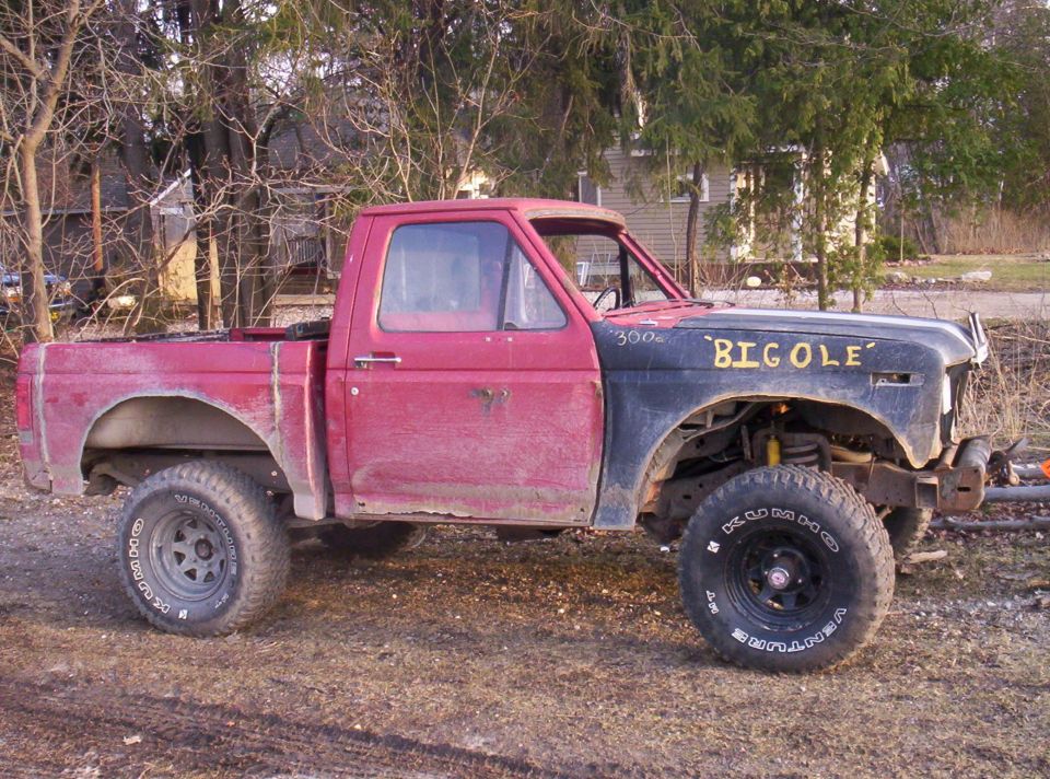 Lifted faded red Ford truck with black front clip painted 'BIG OLE', big Kumho mud tires, parked on dirt