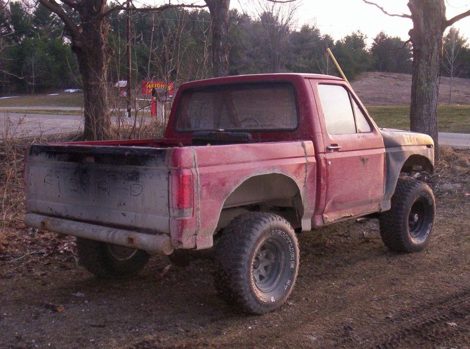 Lifted faded red Ford Bronco with removed rear roof and big off-road tires, seen from rear three-quarter angle by trees