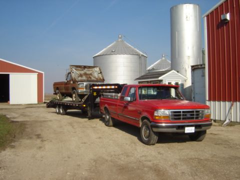 1992 Ford Bronco - A red Ford F-series truck tows a rusty pickup on a trailer, with farm buildings and silos visible.