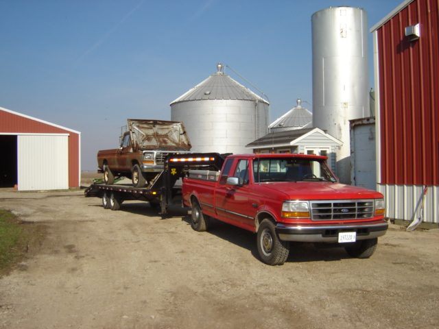 Red Ford F-series truck towing a rusty pickup on a trailer. Farm buildings and silos in background
