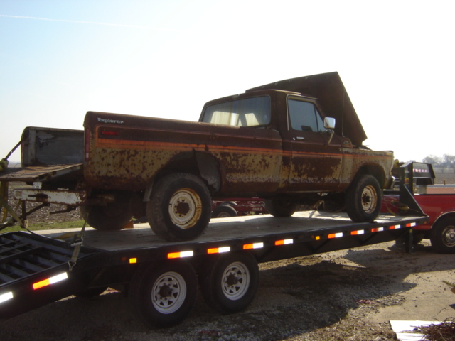 Rusty brown 1970s Ford F-Series pickup truck with orange side trim, hood open, on a flatbed trailer
